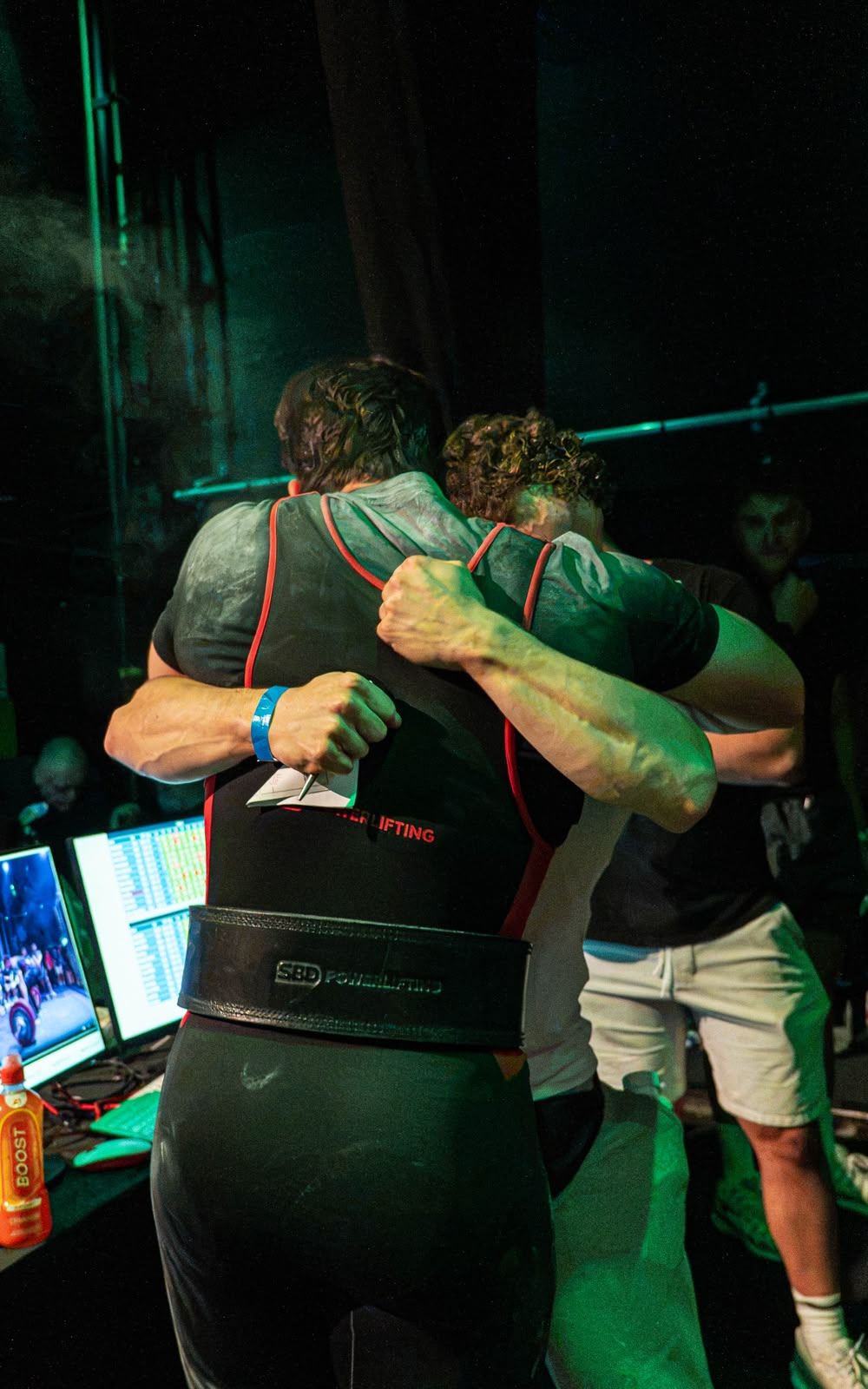 A lifter in a black singlet and belt hugs his coach backstage after a lift