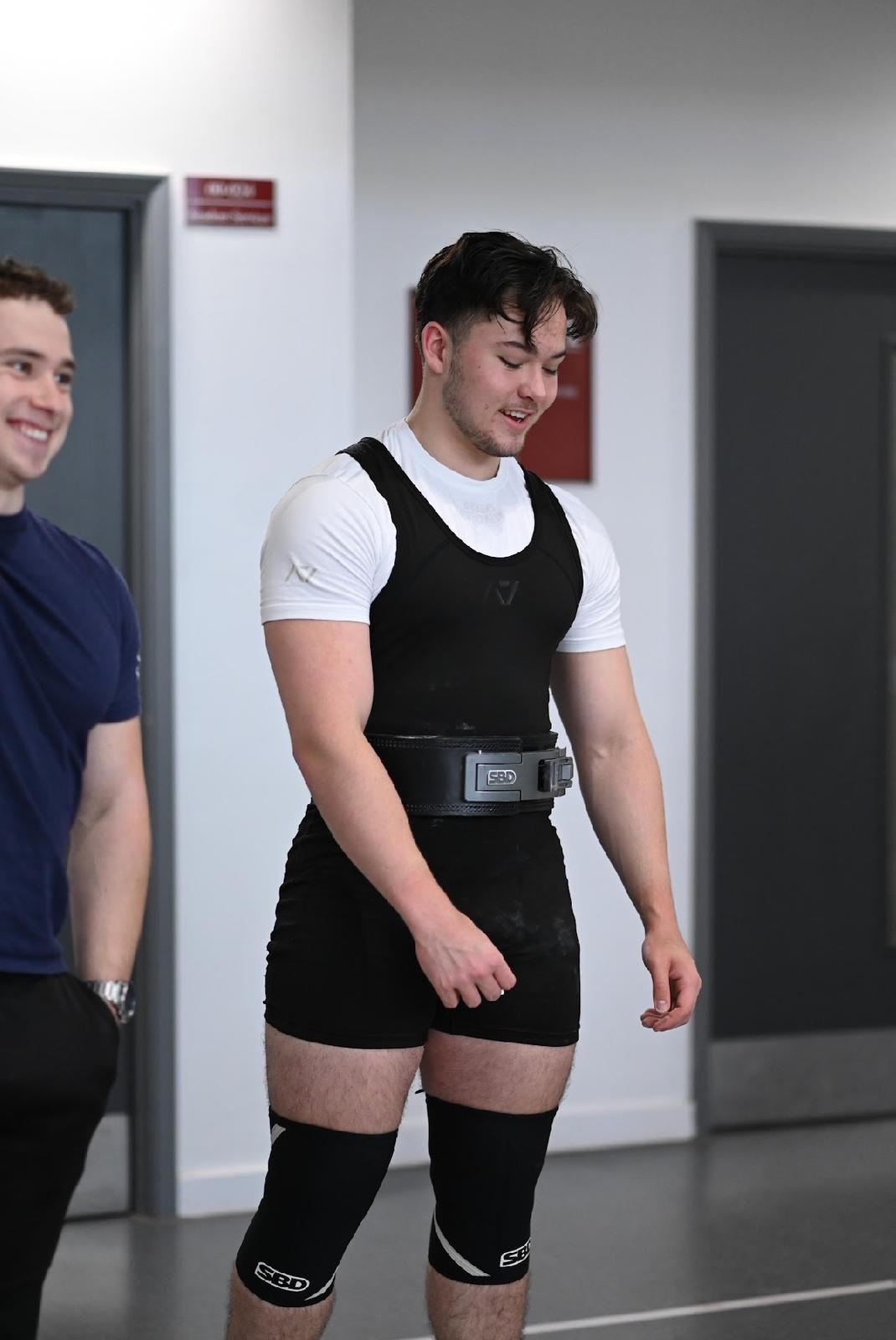 A lifter in a singlet, belt and knee sleeves smiling after a lift