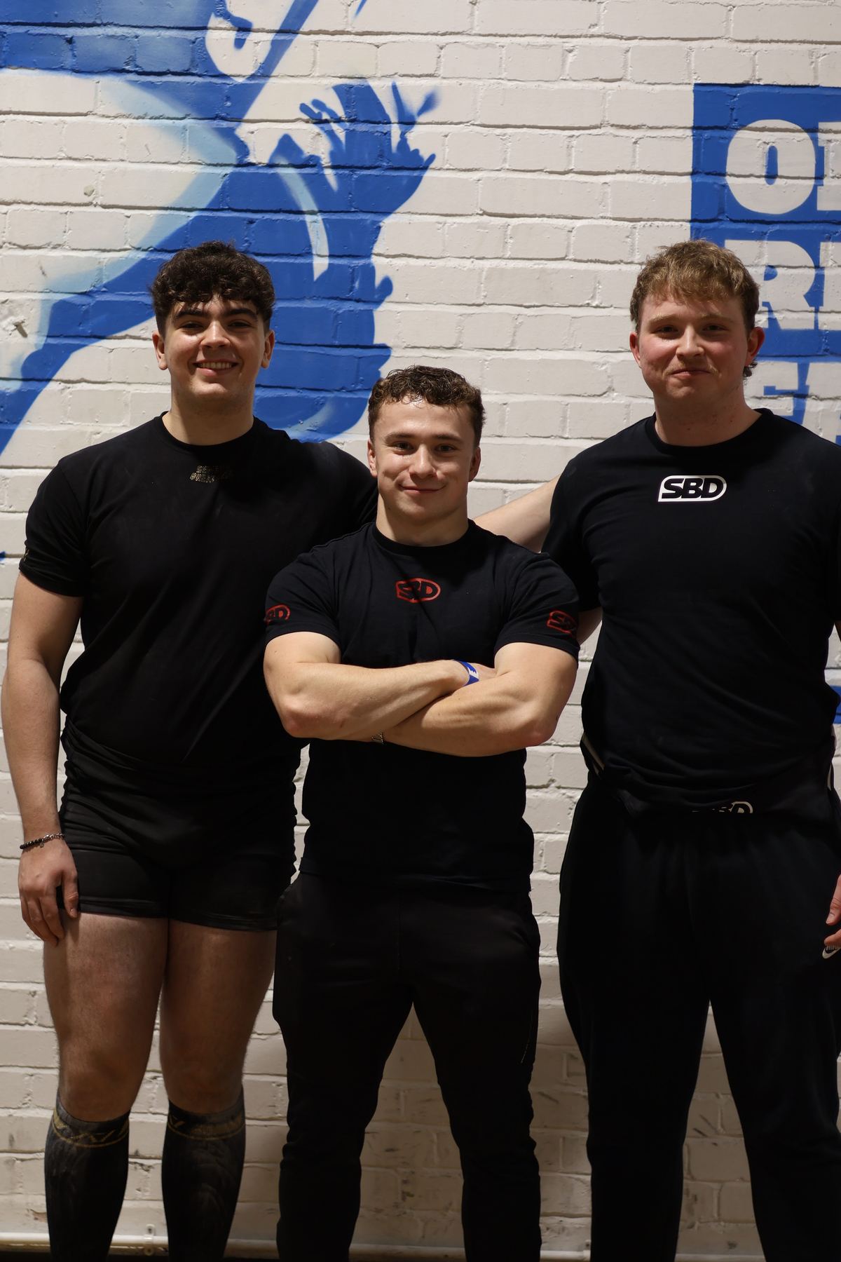 Coach with two lifters in front of a painted brick wall
